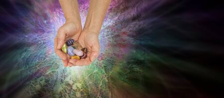 Crystal Healer Offering Chakra Crystals - female therapist holding a set of chakra crystals against a radiating multi-coloured paua shell mosaic background with copy spaceの写真素材