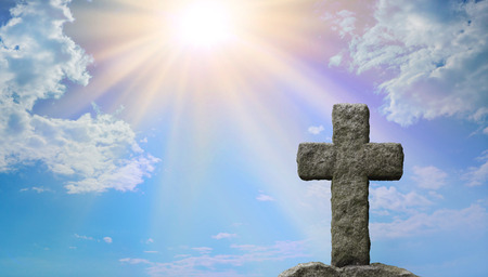 Symoblic Cross of Jesus Christ and God's Light - Rugged stone cross in foreground with a bright blue sky and stuffing sun burst shining down providing copy space on the leftの写真素材
