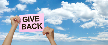 Make someone's day and GIVE BACK - female hands holding high a pink placard that says GIVE BACK against a wide blue sky and fluffy cloud summer backgroundの写真素材