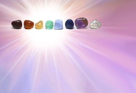 Chakra coloured tumbled healing crystal healing stones template -  bright white light burst on a pink purple graduated background with a row of crystals top left and copy spaceの写真素材