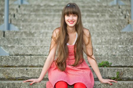 Beautiful girl in a red dress sitting on the gray concrete stepsの写真素材