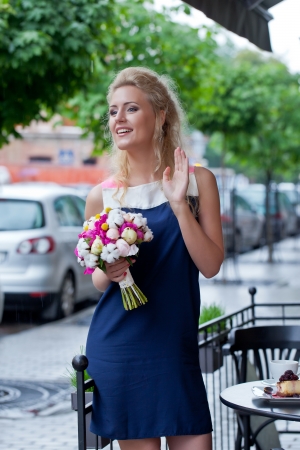 a beautiful young blond girl in summer dress with a bunch of flowers is waving to her friendsの写真素材