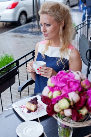 a beautiful young blond girl in summer dress at the table in pavement cafe holding a tea cup is looking thoughtfully in the distanceの写真素材