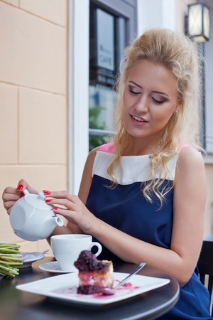 a beautiful young blond girl in summer dress at the table in pavement cafe is going to pour tea from the teapot into the cupの写真素材