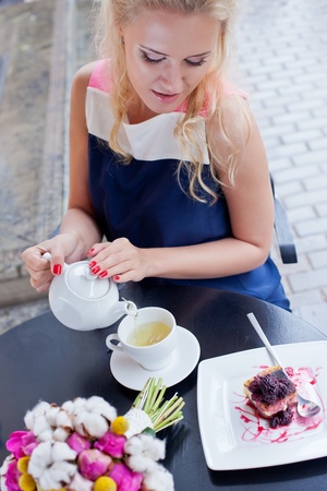 a beautiful young blond girl in summer dress at the table in pavement cafe is pouring green tea from the teapot into a white ceramic cupの写真素材