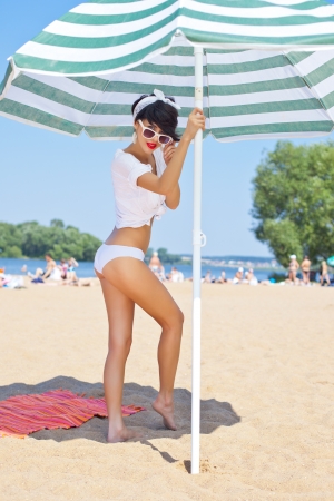a beautiful young girl in retro look with red lips in a white swimsuit, a bandana and sunglasses is standing on the sand under the beach umbrella の写真素材