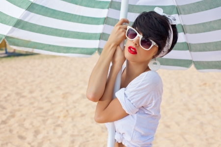 a beautiful young girl in retro look with red lips in a white swimsuit, a bandana and sunglasses is standing under the beach umbrella and is looking into the cameraの写真素材