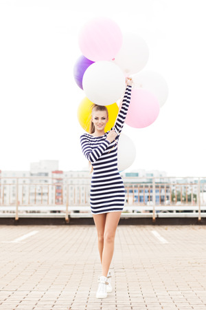 beautiful girl with ponytail hair in short black and white striped dress and white high sneakers walks forward holding bunch of multicolored balloonsの写真素材