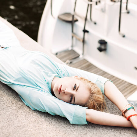 top view of beautiful blonde girl with blue eyes in transparent top lies on pier against white boat and life ringの写真素材
