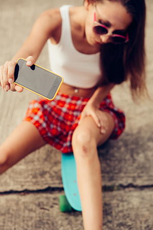 Fit beautiful girl in a red tartan skirt with skateboard taking selfie self portrait of her broken knee at the camera on her smartphone digital camera. Outdoor lifestyle picture on a sunny summer day.の写真素材