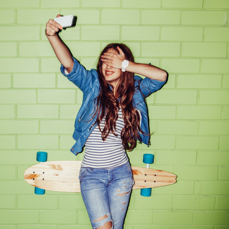 young beautiful long-haired brunette girl with wooden longboard skateboard taking a selfie self portrait of herself coquette at the camera on her smartphone digital camera near the green brick wallの写真素材