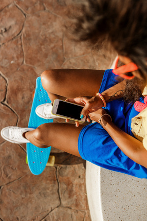 Outdoor lifestyle close-up of black sporty lady in bright outfit and sunglasses. Young hipster girl sitting at skate park and texting on her phone. She is happy. Sunny summer day. Swag, fashion.の写真素材