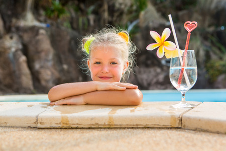 Cute little blonde girl swimming in big pool. Big glass with water, straw and frangipani stay on the pool edge. Little lady smile and look to the camera. Sunbathing and leisure on sunny summer day.の写真素材