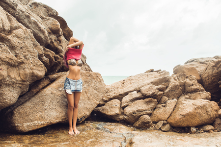 Sexy slim lady in pink t-shirt and denim overall undress on the rock beach sea ocean shore.の写真素材