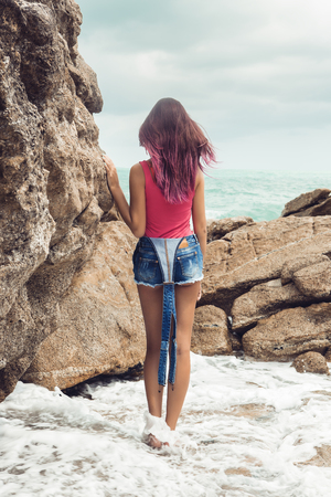 Sexy woman in jeans overall and pink t-shirt stay near big stone at rock beach sea ocean shore. Waves under her feet. View from back. Modern hipster girl have fun. Good sunny summer day for swim.の写真素材