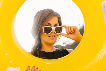 Close up portrait of a beautiful sporty woman in shirt correct her sunglasses and smile through big yellow inflatable ring to the camera. Beauty sunshine cute girl on a sand beach with large stone.の写真素材