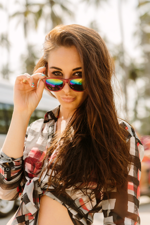 Close up of a young sexy stylish hipster woman on tropic street on a sunny summer day look to the camera and touch her fashion sunglasses. Swag urban teen girl. Outdoor lifestyle portrait.の写真素材