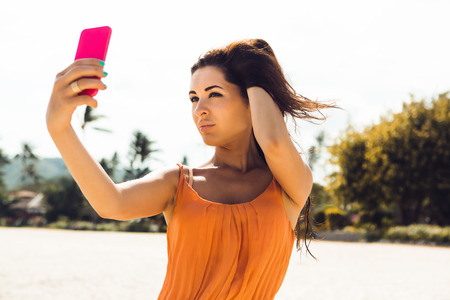 Close up of sexy stylish hipster lady in summer dress, sunglasses make selfie self portrait on a smartphone photo digital camera on a tropical beach. Swag urban teen girl. Outdoor lifestyle portrait.の写真素材
