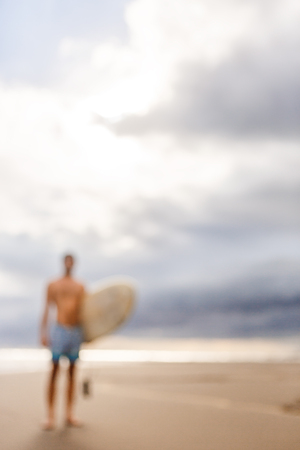 Out of focus, blurry image of handsome man with surfboard stand at sand of surf spot at sea ocean beach after surfing. Concept of sport, freedom, happiness, new modern life, hipster, generation Y.の写真素材