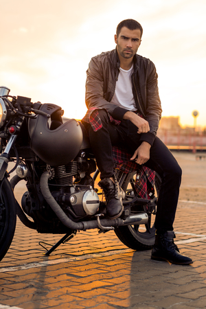 Handsome rider guy with beard and mustache in black leather biker jacket, boot and jeans sit on classic style cafe racer motorcycle. Bike custom made in vintage garage. Brutal fun urban lifestyle.の写真素材