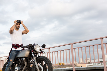 Handsome rider man with beard and mustache posing while put on black moto sunglasses near classic style biker cafe racer motorcycle. Bike custom made in vintage garage. Brutal fun urban lifestyle.の写真素材