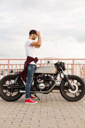 Handsome rider man with beard and mustache put on black moto glasses and ready to go on classic style biker cafe racer motorcycle. Bike custom made in vintage garage. Brutal fun urban lifestyle.の写真素材