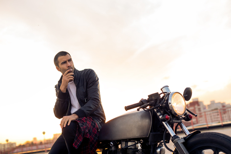 Handsome rider guy with beard and mustache in black biker jacket smoking while sit on classic style cafe racer motorbike at sunset. Bike custom made in vintage garage. Brutal fun urban lifestyle.の写真素材