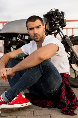 Rider guy with beard and mustache in white t-shirt and red sneakers near his classic style biker cafe racer motorcycle and look forward. Bike custom made in vintage garage. Brutal fun urban lifestyle.の写真素材