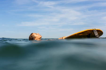 Beautiful sporty surfing girl in sexy bikini swimwear swim near her big longboard surf surfboard board on sunrise or sunset in ocean. Modern active sport lifestyle and summer vacation.の写真素材