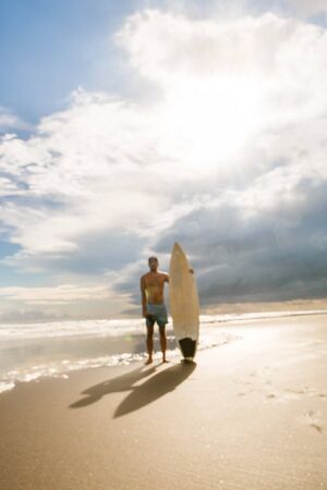 Out of focus, blurry image of handsome man with surfing surf board stand at sand of surf spot at sea ocean beach. Concept of sport, freedom, happiness, new modern life, hipster, generation Y.の写真素材