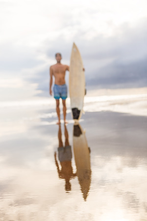 Out of focus, blurry image of handsome man with surfboard stand at sand of surf spot at sea ocean shore after surfing. Concept of sport, freedom, happiness, new modern life, hipster, generation Y.の写真素材