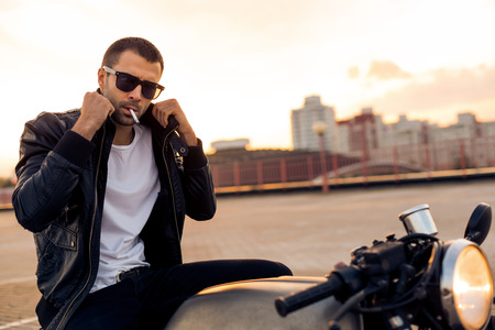 Handsome happy rider man with beard and mustache in black fashion sunglasses smoking cigaret and correct biker jacket sit on classic style cafe racer motorbike at sunset. Brutal fun urban lifestyle.の写真素材