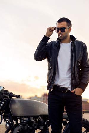 Closeup of a handsome rider guy with beard and mustache in black biker jacket take off fashion sunglasses smoking cigaret near classic style cafe racer motorbike at sunset. Brutal fun urban lifestyle.の写真素材