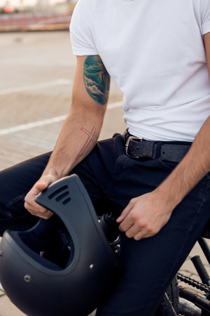 Close up of a handsome rider man in white blank t-shirt with helmet near classic style cafe racer motorcycle at sunset. Bike custom made in vintage garage. Brutal urban lifestyle. Outdoor portrait.の写真素材