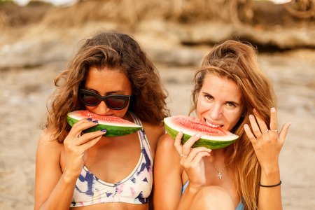 Portrait of a two beautiful surfing girl in sexy bikini eat red sweet juicy watermelon sit on yellow surf longboard surfboard board on sunrise or sunset on a sand beach and look to the camera.の写真素材