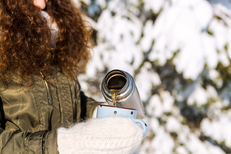 Young beautiful woman in hipster white knitted mittens drink hot tea on a bright winter day. Close-up of a cup filled with hot liquid. Trees in pure first snow on a background.の写真素材