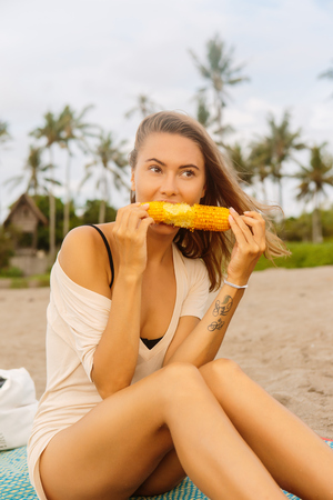 Healthy diet. Vegetarian hipster girl in summer outfit eat fresh organic grilled corn. Sexy lady on sea beach sunset or ocean sunrise. Travel, explore, active yoga and meditation lifestyle concept.の写真素材