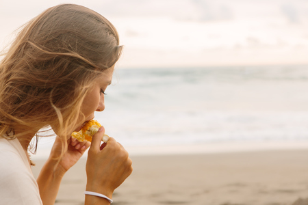 Healthy diet. Vegetarian hipster woman eat fresh organic grilled corn. Sporty lady on sea beach sunset or ocean sunrise. Travel, explore, active, yoga, vegan and meditation lifestyle, dieting concept.の写真素材