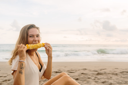 Healthy diet. Vegetarian hipster woman eat fresh organic grilled corn and look to camera. Sporty lady on sea beach sunset or ocean sunrise. Travel, active, yoga, vegan and dieting lifestyle concept.の写真素材