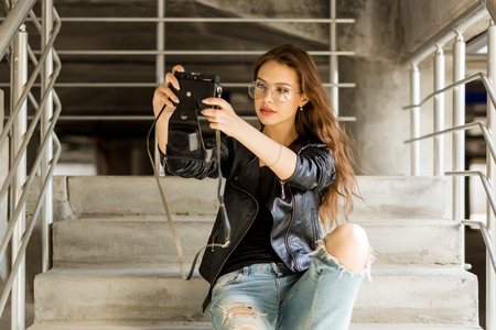 Photo of young cheerful woman in fashion transparent glasses wearing a rock black style leather jacket making selfie photo on vintage camera. Hipster urban style girl. Lifestyle outdoor portrait.の写真素材