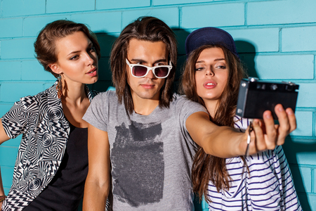 Beautiful best friend hipsters wearing stylish bright outfits and having great time. Handsome boy with two beautiful ladies taking selfie self portrait photo with film camera in front of blue brick.の写真素材