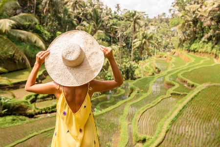 Beautiful young lady in shine through dress touch straw hat. Girl walk at typical Asian hillside with rice farming, mountain shape green cascade rice field terraces paddies. Ubud, Bali, Indonesia.の写真素材
