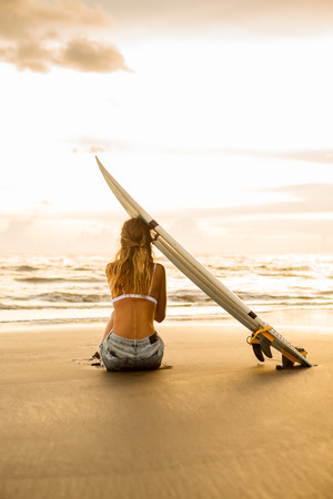 Young surfer woman in sexy white bikini with blank surfing shortboard sit on sand at sunset ocean. Modern family lifestyle, people water sport adventure camp and extreme swim on summer vacation.の写真素材