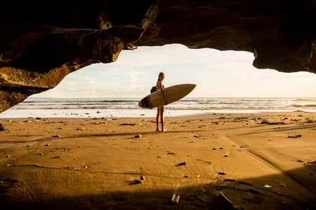 Young surfer woman with surf board pose in cave at beach. Garbage and plastic on sand. Concept of...