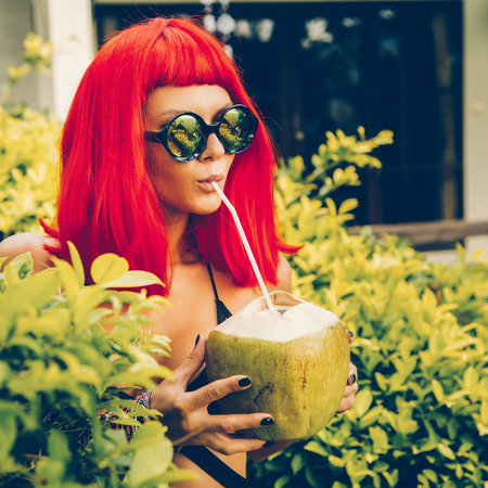 Close up portrait of a sexy thirsty fit woman in red wig, black sensual swimsuit and fashion sunglasses drink coconut water with straw. Beauty cute girl on a tropical resort. Outdoor summer lifestyle.の写真素材
