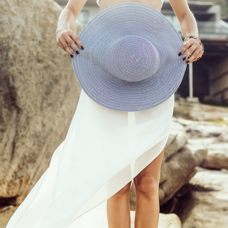 Close up of legs of a beautiful woman stand on rock in white skirt hold her summer straw stripped hat. Beauty cute girl on a tropical beach sea ocean shore with large stones. Outdoor summer lifestyle.の写真素材