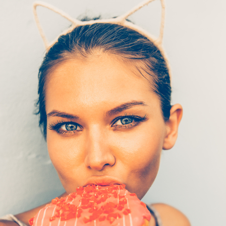 Woman enjoy tasty sweet heart shaped donut and look to camera. Beauty cute girl in a tropical resort. Outdoor lifestyle portrait on hot sunny summer day. Concept of love, feelings, positive emotions.の写真素材