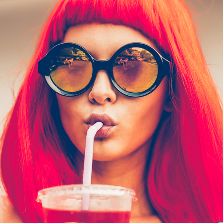 Close up portrait of a beautiful thirsty woman in red wig, black sexy swimsuit and fashion sunglasses drink red soda water with straw. Beauty cute girl on a tropical resort. Outdoor summer lifestyle.の写真素材