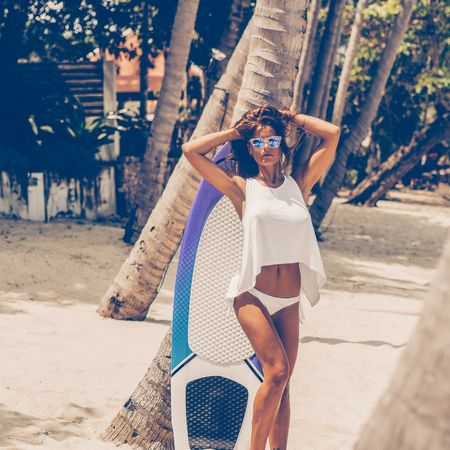 Sexy lady in sensual white tunica on a tropical beach posing with her surfboard between palm trees....