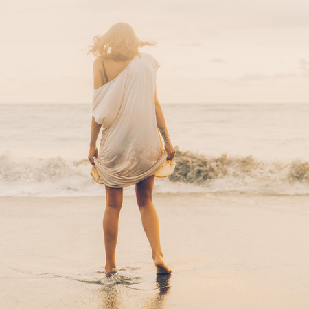 Hipster trendy woman hold her long casual summer dress and walk in little waves on the coastline. Sporty lady on sea sand beach sunset or ocean sunrise. Travel, active, yoga lifestyle concept.の写真素材